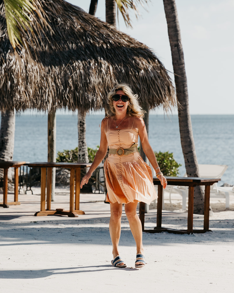 Woman in a peach dress standing on a beach with palm trees and a thatched roof structure in the background.