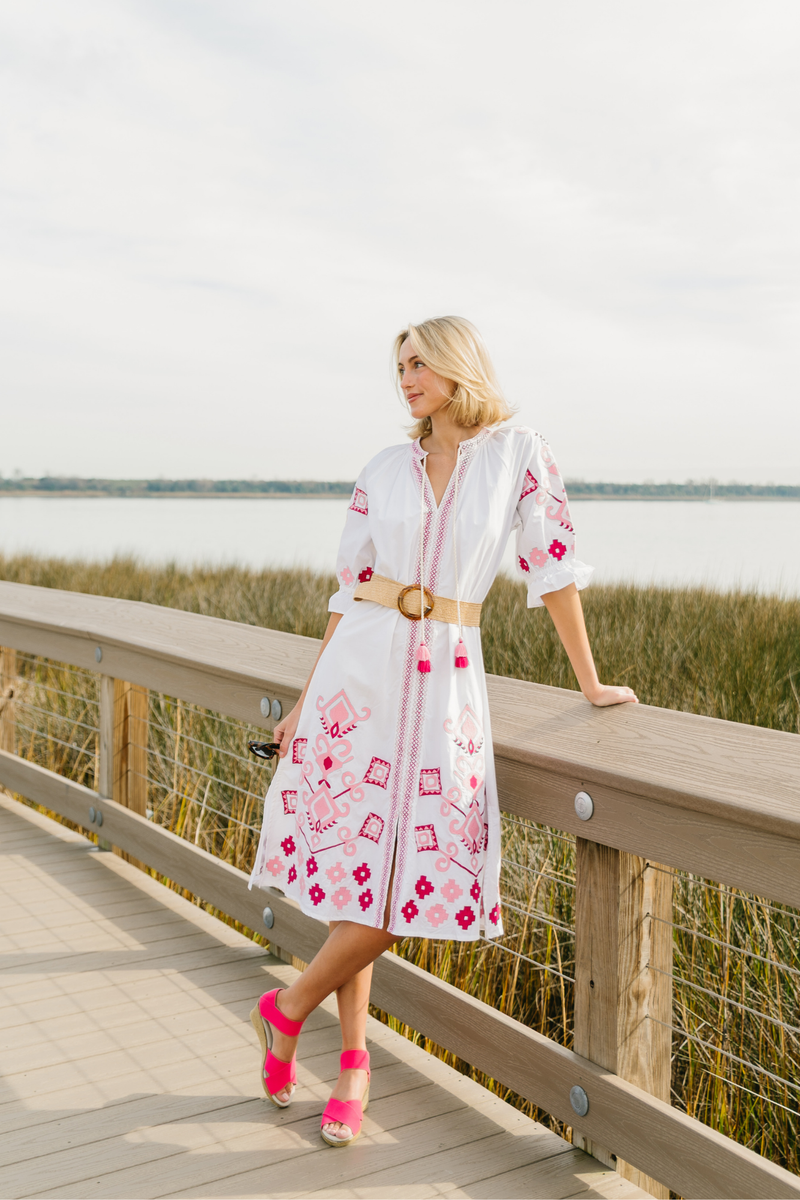 Woman in a white dress with pink floral patterns standing on a wooden boardwalk by a body of water.